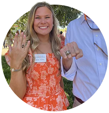 A female student in an orange floral dress holds up her hand to show the Clemson Ring on her ring finger.