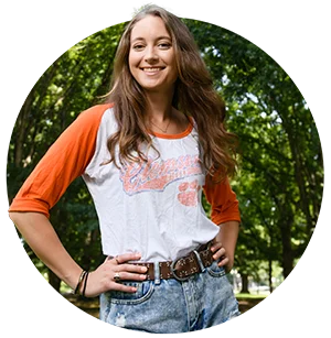 A female student in a faded Clemson shirt and jeans poses and smiles in front of nearby trees.