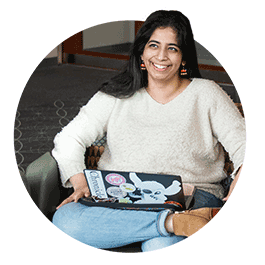Student Shaoni Dasgupta, wearing a white sweater and jeans, sits on a chair in a student lounge with her laptop on her lap.