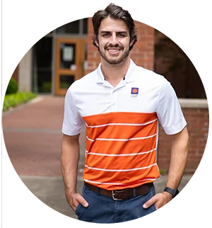 A male student wearing a collared white-and-orange Clemson shirt smiles as he poses in front of an academic building.