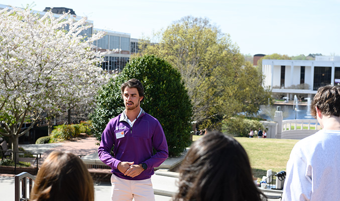 Student tour guide stands on a bench beside Lee Hall and addresses his tour group.