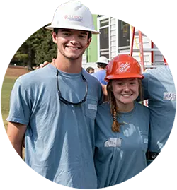 A male student wearing a blue T-shirt and a white hardhat and a female student wearing a blue T-shirt and an orange hardhat stand in front of a Habitat for Humanity home build. 