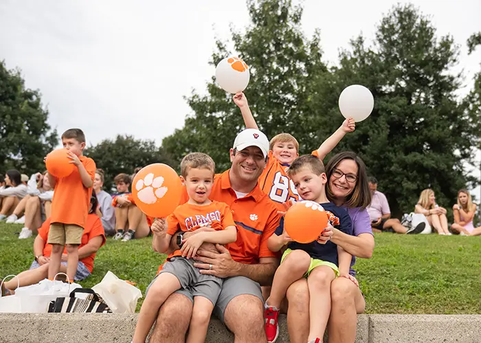 A man and a woman with three young boys, all wearing Clemson spirit gear, sit on a concrete ledge during the First Friday Parade. The young boys are holding orange and white balloons adorned with Clemson Tiger paws.