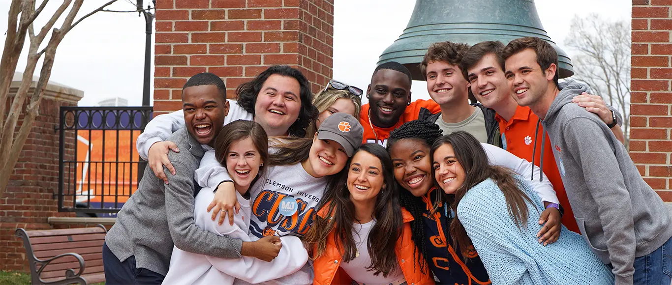 A dozen students are huddled together with their arms around each other, smiling for a group photo in front of the brick Class of 1939 bell tower on Clemson’s campus 