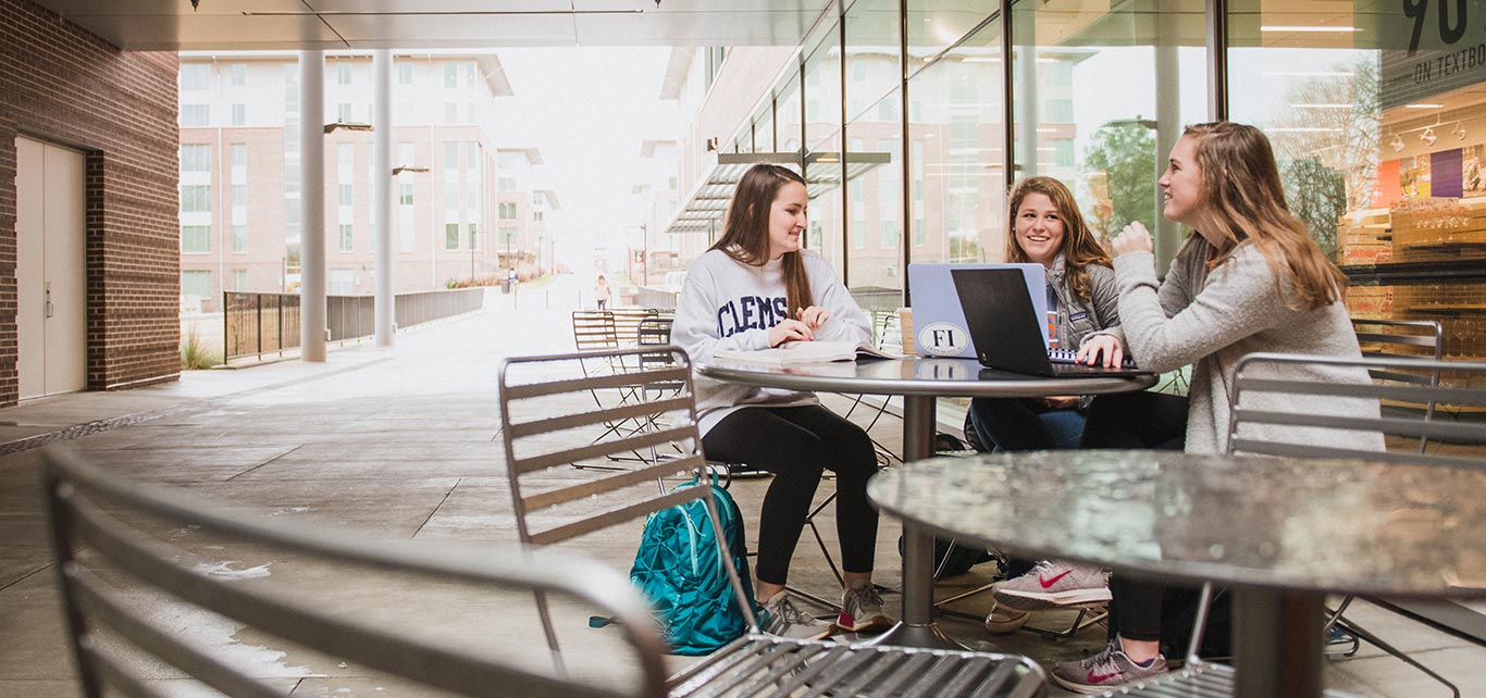 Three female students sit together at an outdoor table. Two female students on the right have their laptops open, while the female student on the left has a textbook open. 