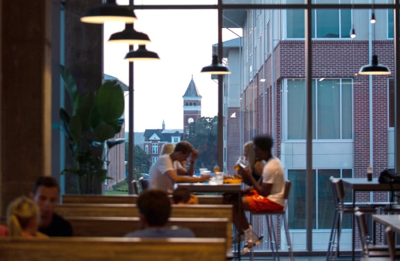 Clemson students sit together in a dining hall in Douthit Hills as the sun sets and highlights Tillman Hall.