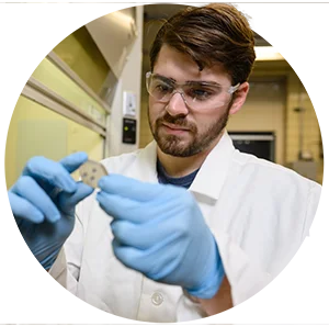 A male student in a white lab coat and safety goggles inspects a piece of glass he holds in his gloved hands.