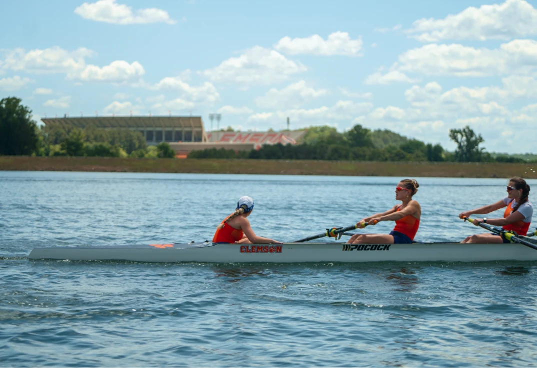 Julia Walsch leads her team in a rowing shell on Lake Hartwell with Memorial Stadium in the background.