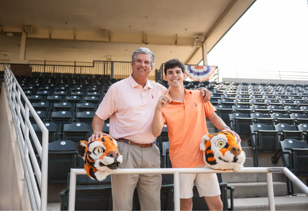 A man wearing khaki pants and a light orange polo shirt has his arm around his son, who wears khaki shorts and an orange polo shirt. They are standing among seats in a baseball stadium, and both pose with a head of a Clemson Tiger mascot
