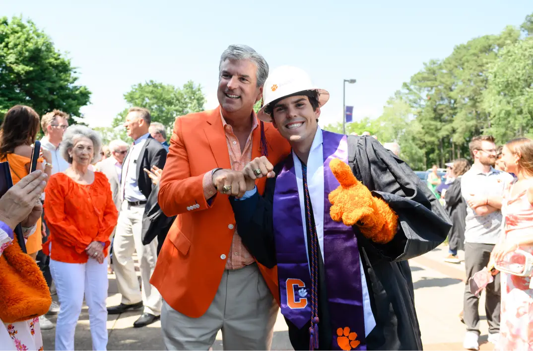 A man wearing khaki pants, an orange windowpane-patterned button-down shirt and an orange blazer holds up his Clemson Ring next to his son, who wears a black graduation gown, purple graduation stole and white construction hard hat. On one of his hands, he wears a fuzzy orange Clemson Tiger mascot sleeve, and on the other, he holds up his Clemson Ring.