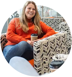 A female student wearing jeans and an orange sweater smiles while sitting in a chair and holding a cup of coffee.