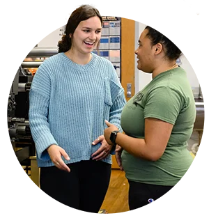 A female student in a blue sweater laughs as she speaks with a female student in a green shirt.