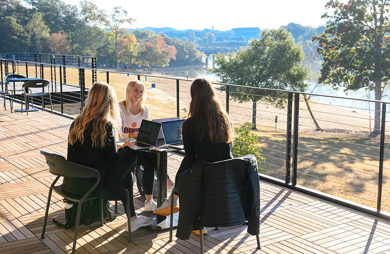 Three female students sit together at a table on an autumn day with their laptops open in an outdoor space featuring a view of Lake Hartwell and a sandy beach.