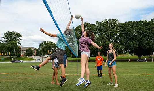 A group of six students plays volleyball on Bowman Field. A female student attempts to tap the ball over the net while a male student tries to block the attempt.