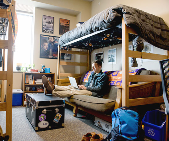 Lights streams in the window as a male student sits on a futon under his lofted bed in an on-campus dorm, using his laptop.
