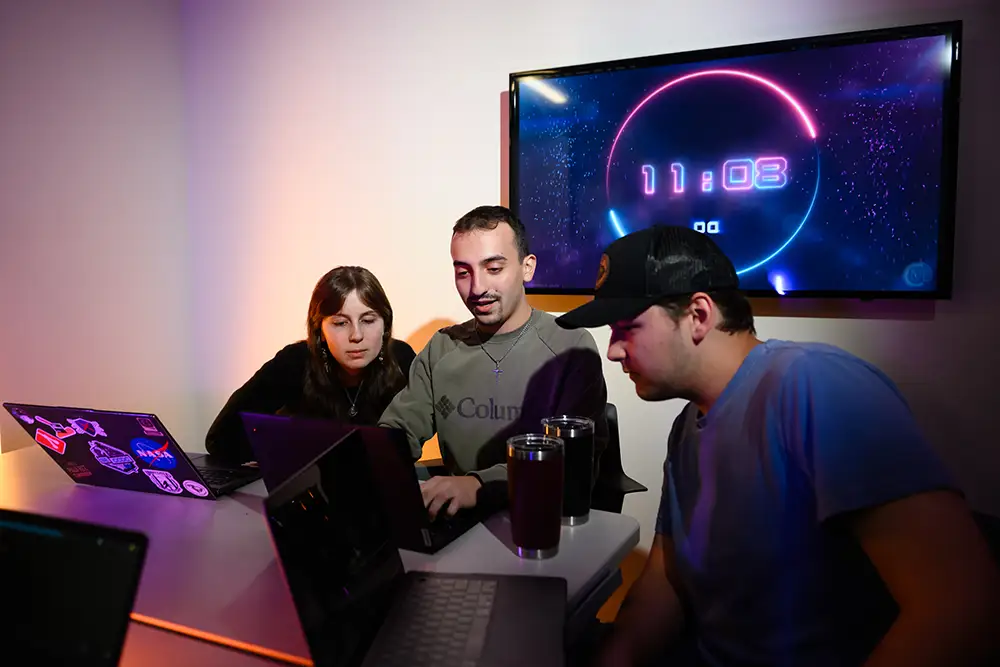 Two male students and one female student sit closely at a table as they look at three laptop screens on a table in a room that is dimly lit with colorful accent lighting and a screen with a countdown clock that reads 11:08.