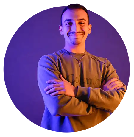 A male student smiles and stands with his arms crossed against a purple-lit studio background that casts soft blue and orange tones across his face and clothing. He wears a sweatshirt and cross pendant necklace.