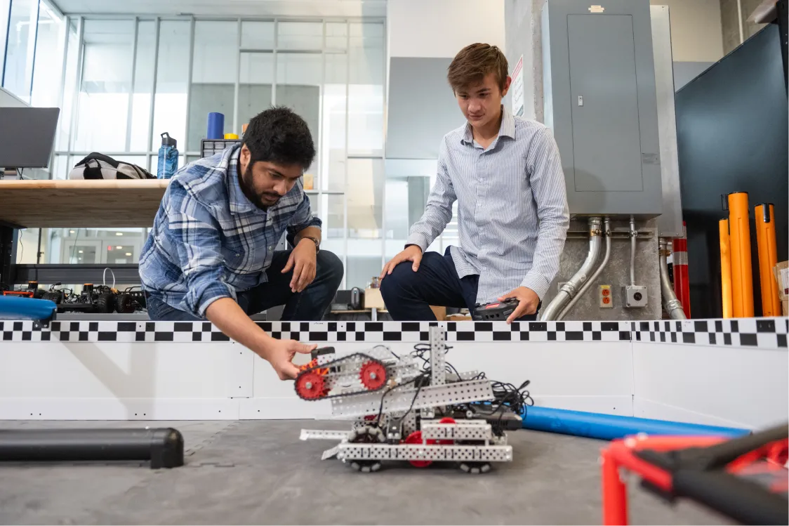Two male students wearing blue jeans and long-sleeved button-down shirts lean over a track wall and examine a small robot.