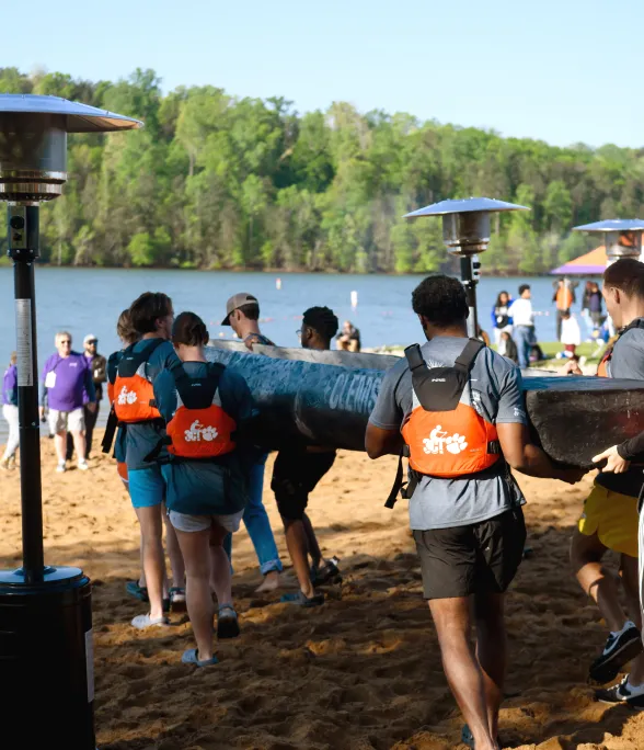 A group of students walks along a sandy shore on a lakeside beach while carrying a blue can