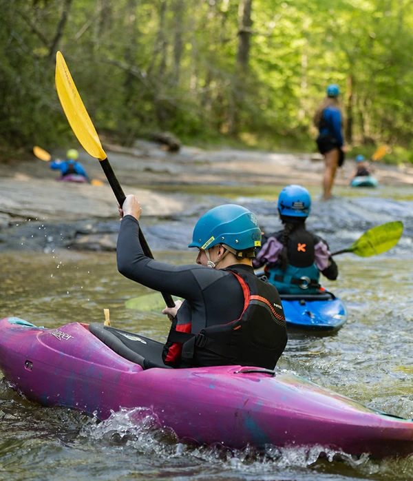 A group of leisure skills students kayaks on whitewater rapids in a river, with a student in a purple kayak and black wetsuit in the foreground.