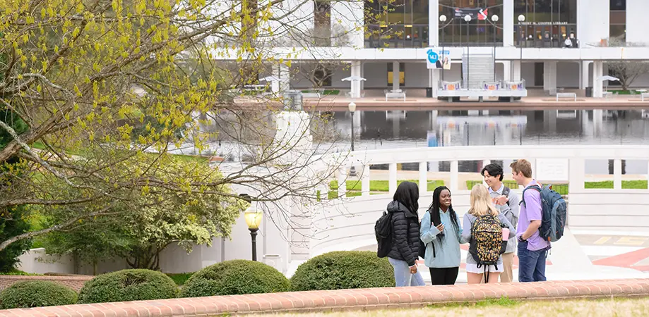 Three female students and two male students gather on a sidewalk on campus in front of the Amphitheater and the Cooper Library Reflection Pond.