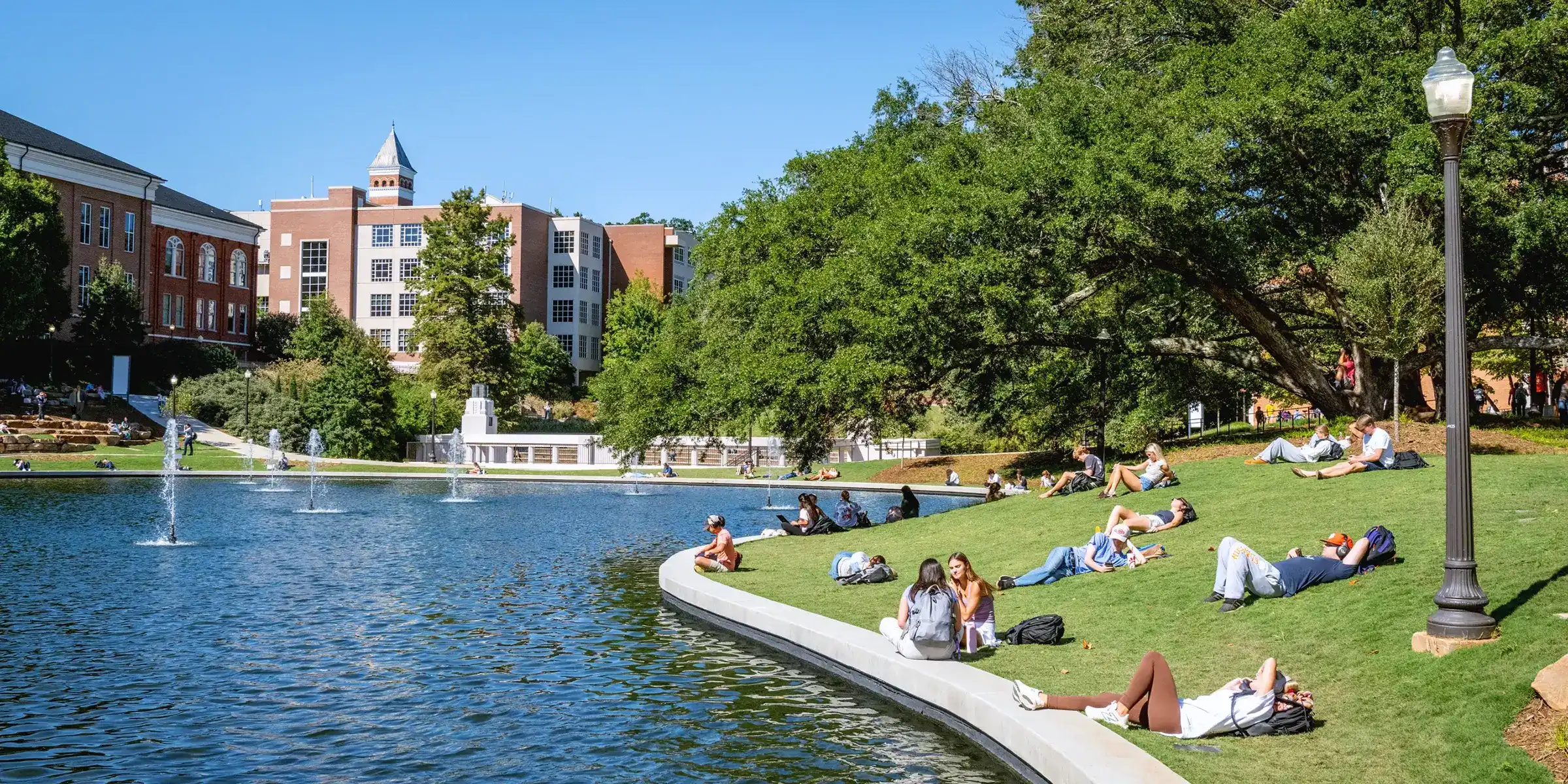 Several college students lounge on a sunny day on a green space beside a campus pond with fountains, surrounded by trees and brick academic buildings. 