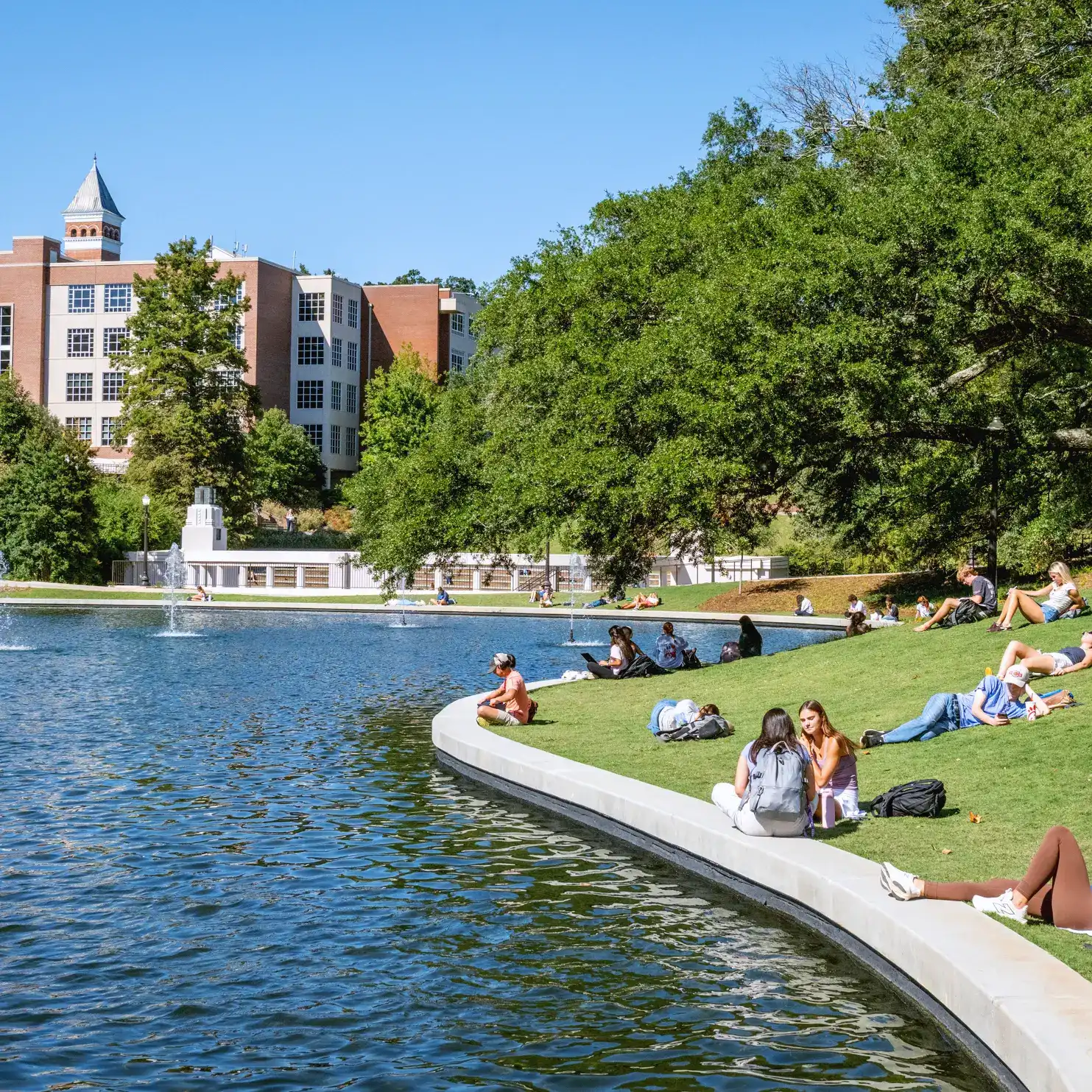 Several college students lounge on a sunny day on a green space beside a campus pond with fountains, surrounded by trees and brick academic buildings. 