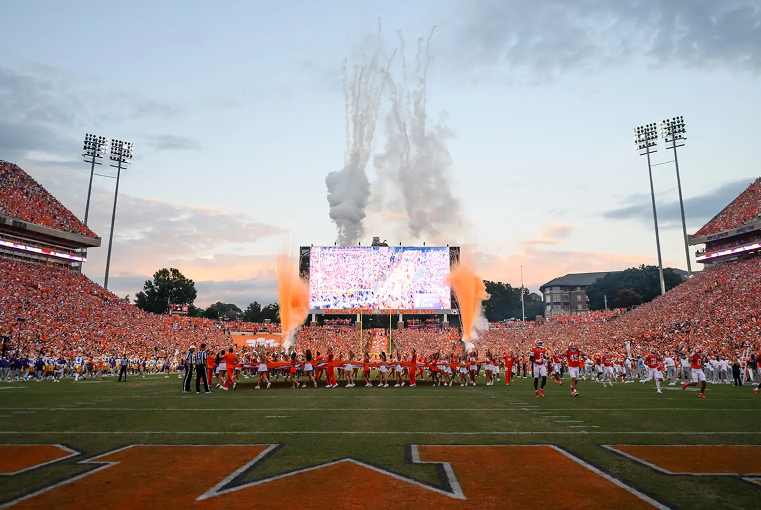 Football players run onto the Memorial Field as celebratory fireworks erupt, with a packed stadium and a large screen in the background.