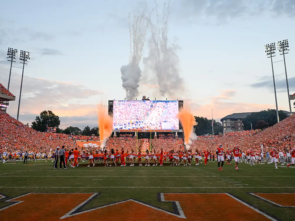Football players run onto the Memorial Field as celebratory fireworks erupt, with a packed stadium and a large screen in the background.