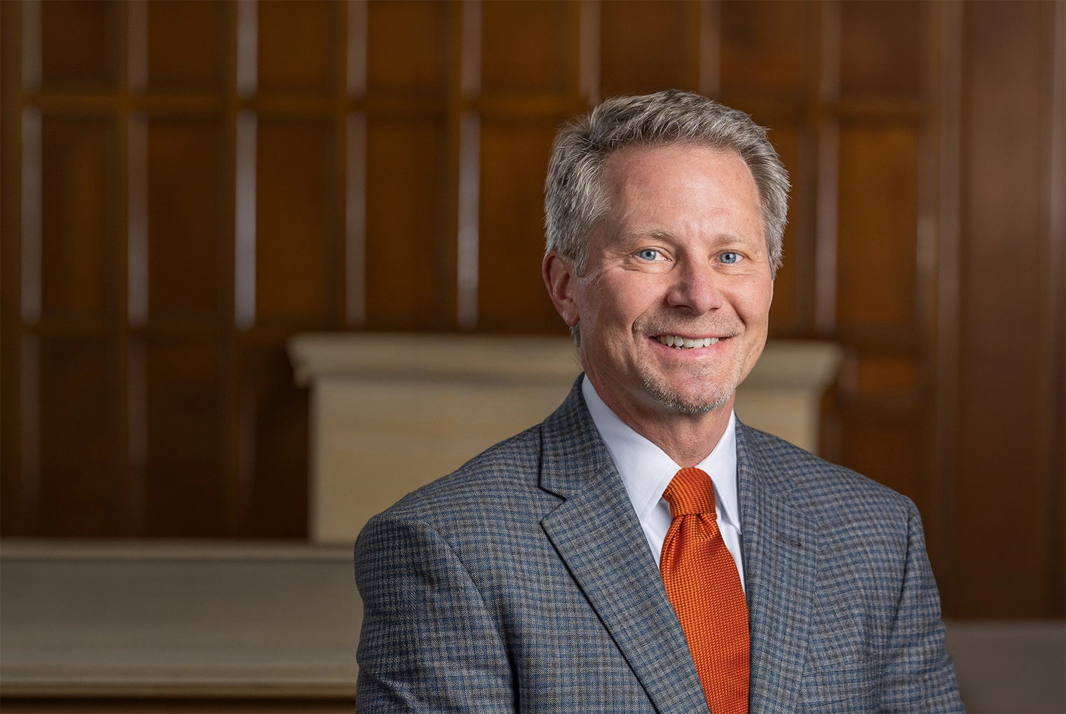 A smiling President-Elect Guskiewicz in a gray suit with an orange tie in an indoor office setting.