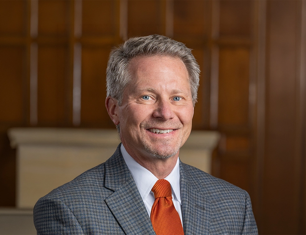 A smiling President-Elect Guskiewicz in a gray suit with an orange tie in an indoor office setting.