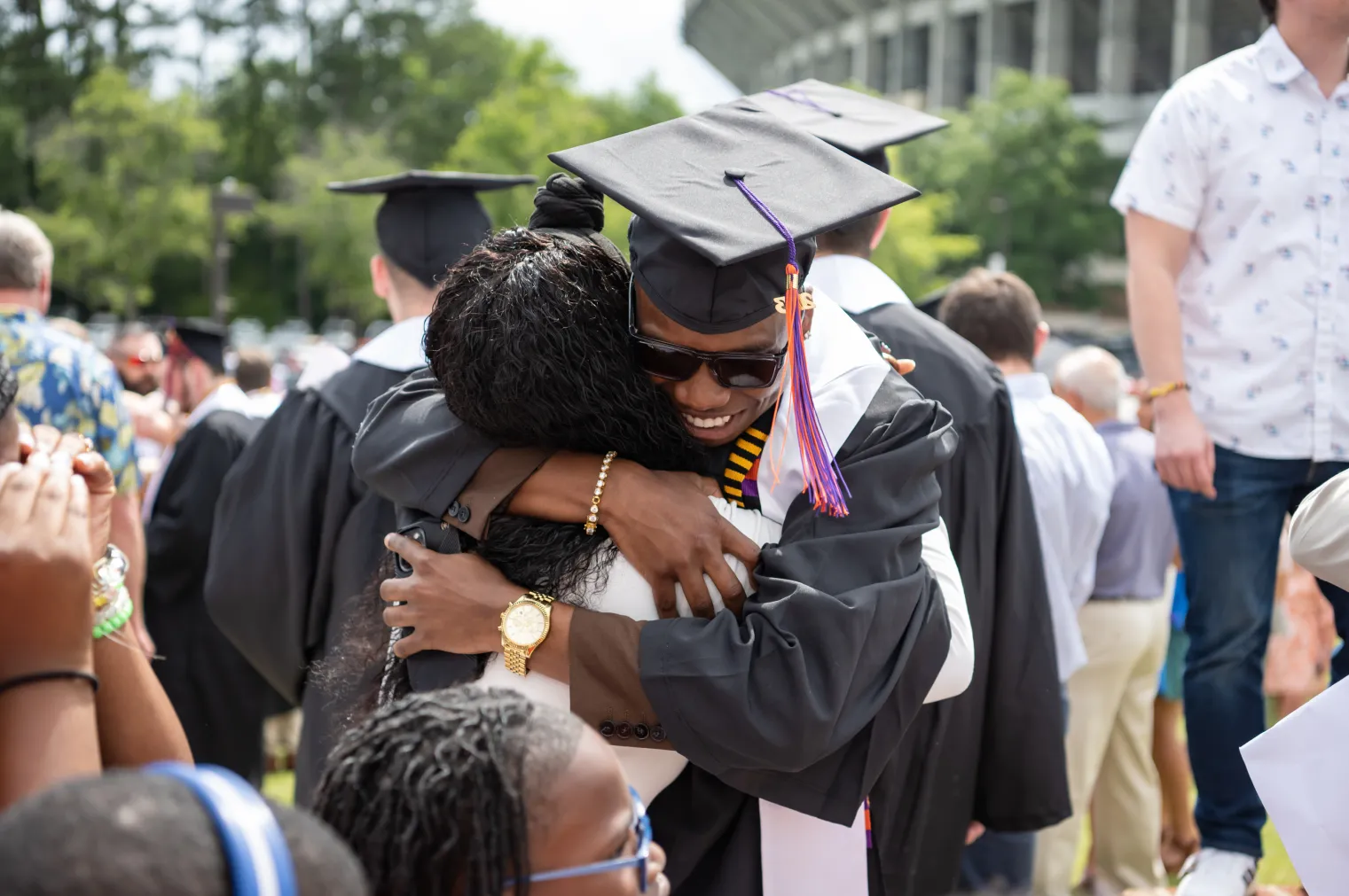 Two new Clemson Graduates wearing their caps and gowns, on the lawn of littlejohn colosseum, embrace in a hug after their commencement ceremony