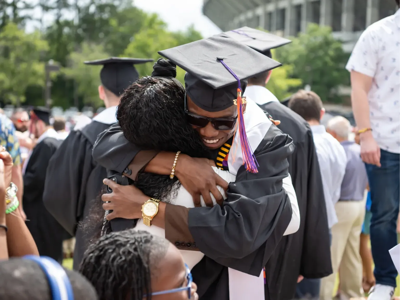 Two new Clemson Graduates wearing their caps and gowns, on the lawn of littlejohn colosseum, embrace in a hug after their commencement ceremony