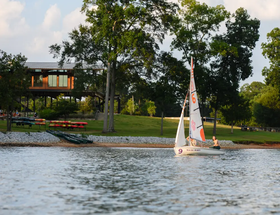 A female student pilots a small Clemson branded sailboat on Lake Hartwell in front of the Andy Quattlebaum Outdoor Center