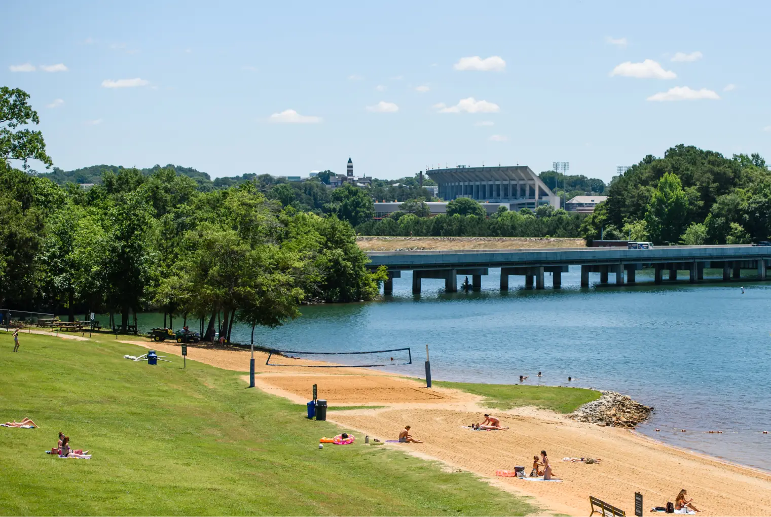 Students lounge on a large green lawn transitioning into a sandy beach on the shore of Lake Hartwell. Clemson's campus is visible in the background.