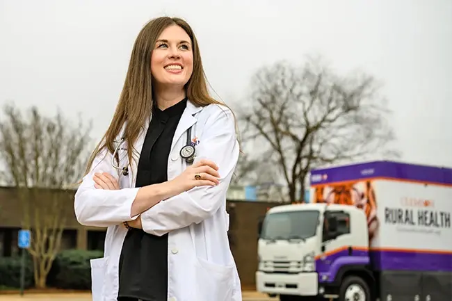 A woman wearing a black top and a white lab coat with a stethoscope around her neck looks off into the distance while standing in front of a purple, orange and white Clemson Rural Health mobile clinic truck.