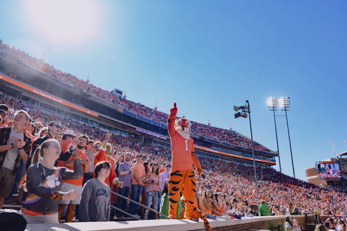 The Clemson Tiger mascot stands on top of a concrete wall with a finger pointed in the air next to stands in Memorial Stadium, which are filled with Clemson students and fans on a sunny football game day. 