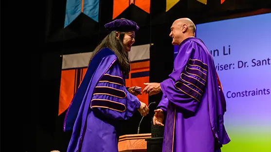 University President Jim Clements congratulates a female student on stage at the doctoral hooding ceremony. Both are wearing purple academic regalia.