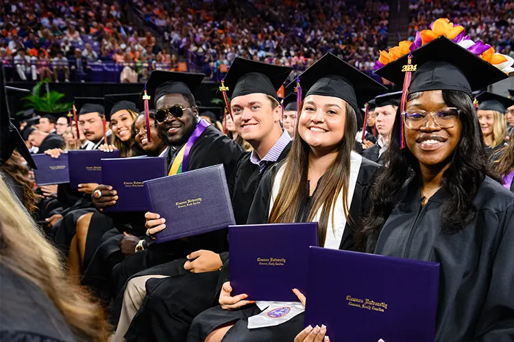 Six undergraduate students — three male and three female — sit in a row during a graduation
ceremony and hold up their diploma covers. All wear black graduation gowns and caps.