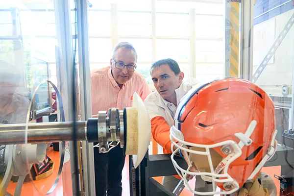 Two male professors observe a football helmet being tested in a lab. The helmet is secured on a mannequin head while a padded device prepares to simulate impact against it.