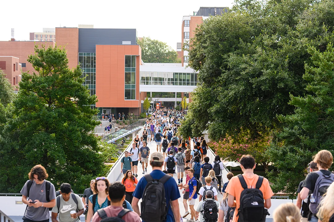 A large crowd of students carrying backpacks walks down stairs and across a pedestrian
bridge with academic buildings constructed from brick and glass in the background.