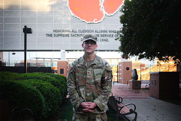 A male student in a military camouflage outfit wearing glasses stands in front of Memorial Stadium on Clemson’s campus with the orange Tiger Paw and military dedication inscription partially visible.