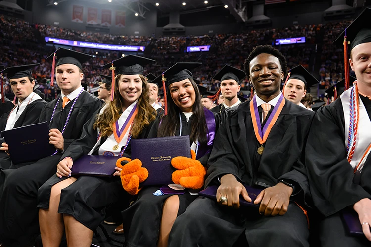 A group of Clemson University graduates in caps and gowns sit in rows during a commencement ceremony and hold their diplomas. The graduate in the center is wearing orange Tiger mascot gloves.