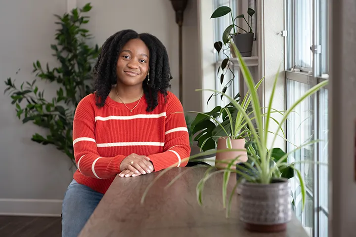 A female student wearing denim jeans and a red sweater with white stripes stands next to a shelf with two green potted plants sitting on it.