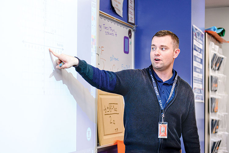A male student teacher wearing a navy polo shirt and jacket stands at the front of a high school classroom and points to a math problem projected on a screen.