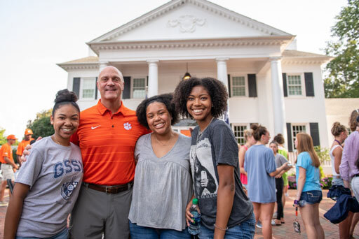 Three Black female students smile with President Clements on the front lawn of the President's Home during the freshman BBQ.