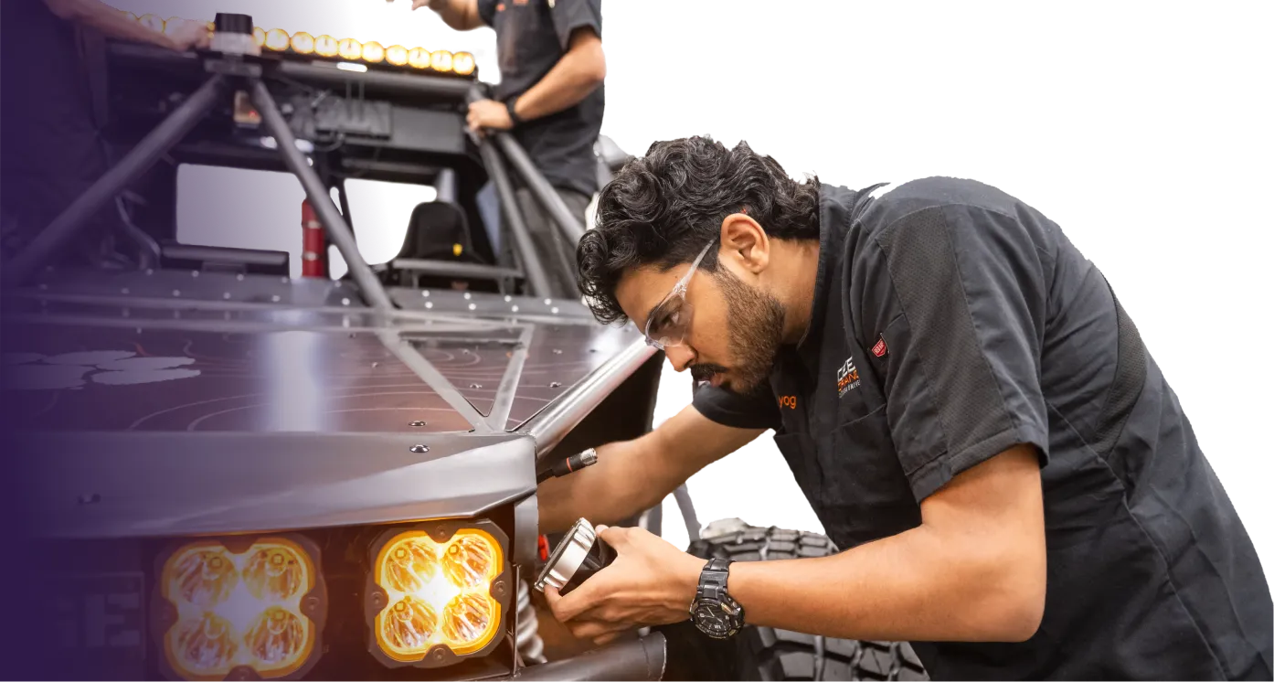 A male student working in an engineering prototyping lab wears safety glasses as he adjusts a light feature on a vehicle frame, with a row of amber lights in the foreground. 