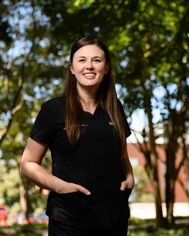 Caitlin Kickham stands with her hands in the pockets of her medical uniform under shade trees on a sunny day