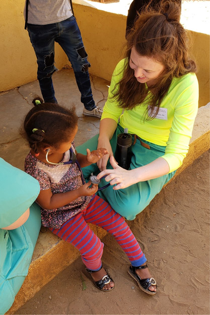 Caitlin sits on a concrete stoop with a child who is wearing a stethoscope. Caitlin points to the diaphram.