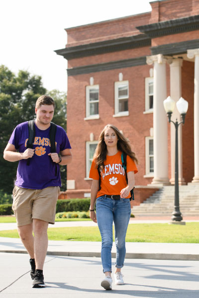 Emily walks and talks with a male Clemson student in front of Sikes Hall. Both wear Clemson shirts and backpacks. 